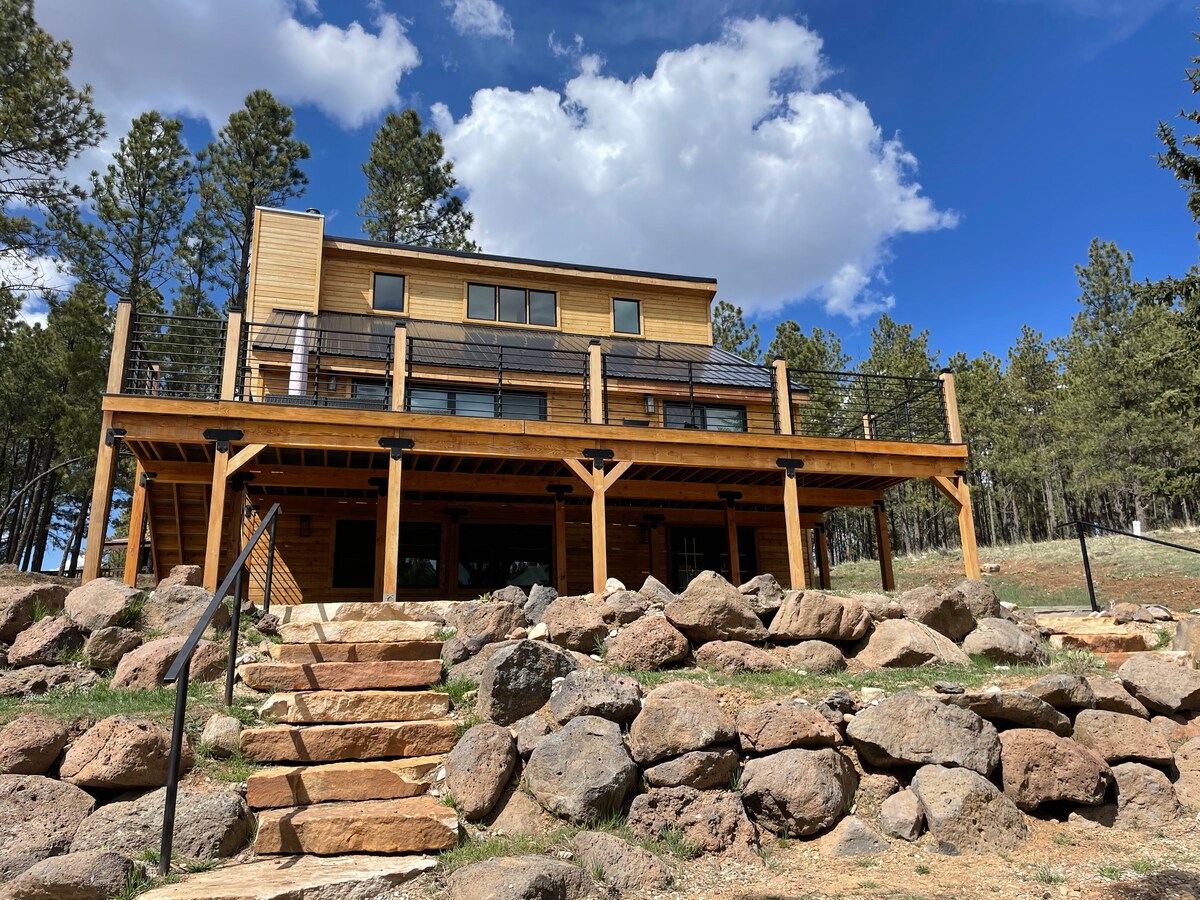 The exterior of the house features a large deck supported by wooden posts, accessible by a stone staircase surrounded by natural landscaping. Tall trees dot the background under a bright blue sky with scattered clouds, creating a harmonious blending of architecture and nature.