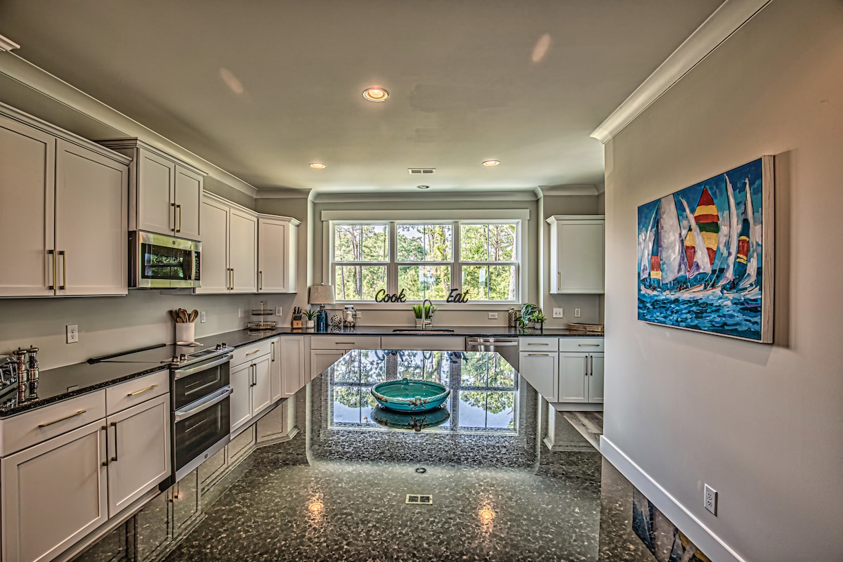 A spacious kitchen features modern appliances and ample counter space, with a central island that serves as a focal point. Large windows allow natural light to fill the room, offering views of the tranquil outdoor surroundings. Light-colored cabinetry complements the sleek design.