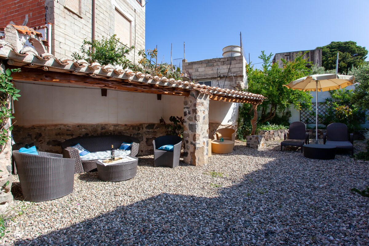 An outdoor courtyard features a shaded seating area with wicker furniture, complemented by cushions. A sun umbrella provides shelter, while lounge chairs invite relaxation. The space is surrounded by greenery and has a gravel floor, offering ample room for outdoor gatherings.