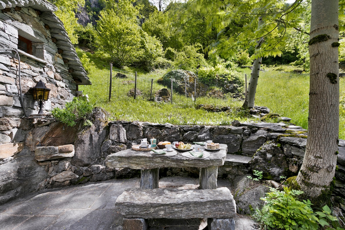 An outdoor stone dining table is set with plates, cups, and utensils, surrounded by lush greenery. The rustic seating area features a backdrop of natural stone and views of a grassy landscape, offering a serene atmosphere for al fresco meals.