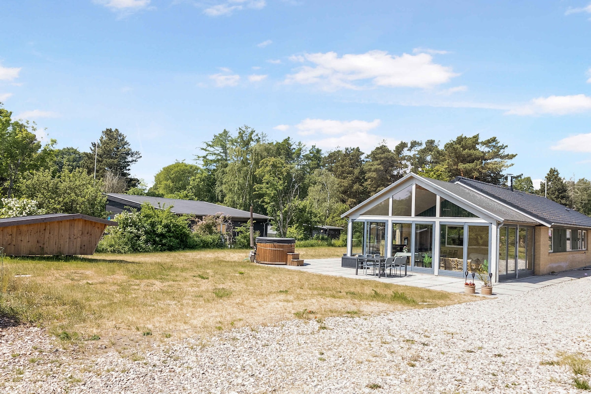 The exterior features a modern summer house, set against a backdrop of lush greenery. A spacious patio is visible, with outdoor seating and a hot tub, surrounded by open grass. Two additional structures are partially hidden among the trees, enhancing the tranquil setting.