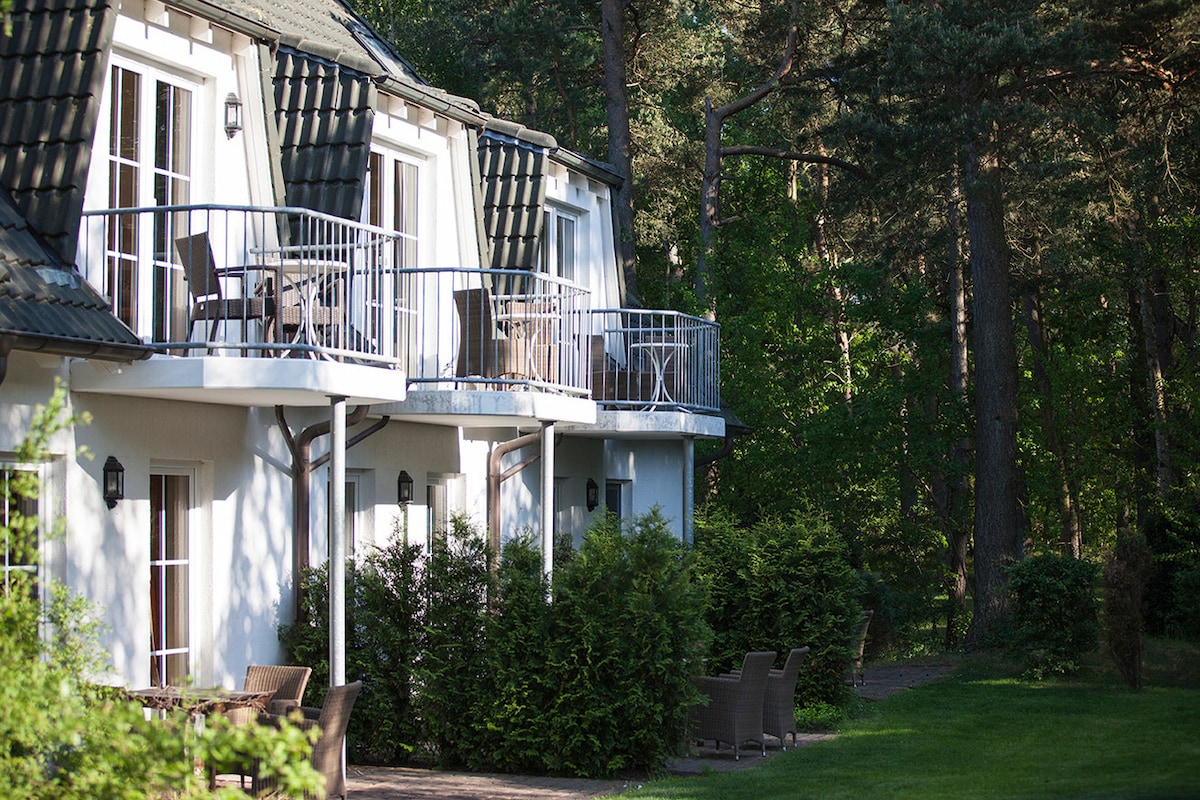 Multiple accommodations are visible, each featuring a private balcony with seating. The light-colored buildings are surrounded by lush greenery and trees, creating a serene retreat atmosphere. Pathways lead through well-kept greenery, enhancing the connection to nature.