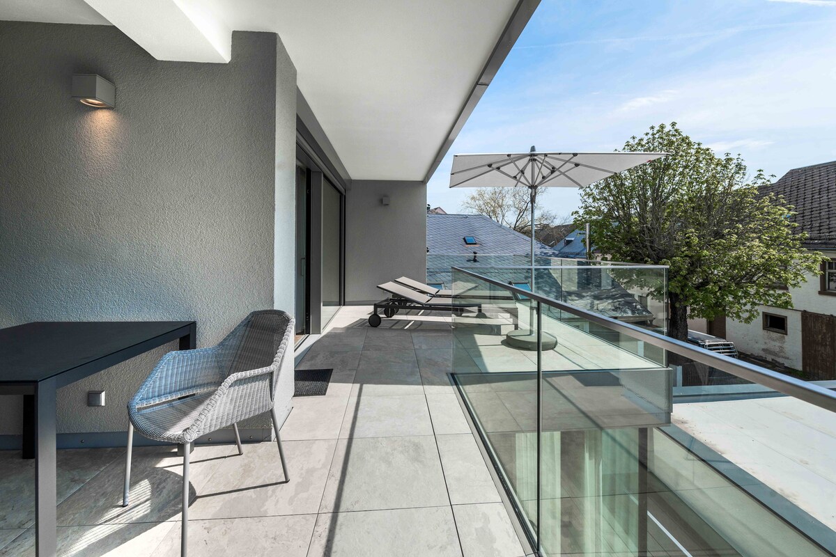 A spacious sun terrace features a table and two sun loungers beneath a parasol. Glass railings provide unobstructed views of the surroundings, with tiled flooring enhancing the outdoor experience. Natural light floods the area, creating a bright and open atmosphere.