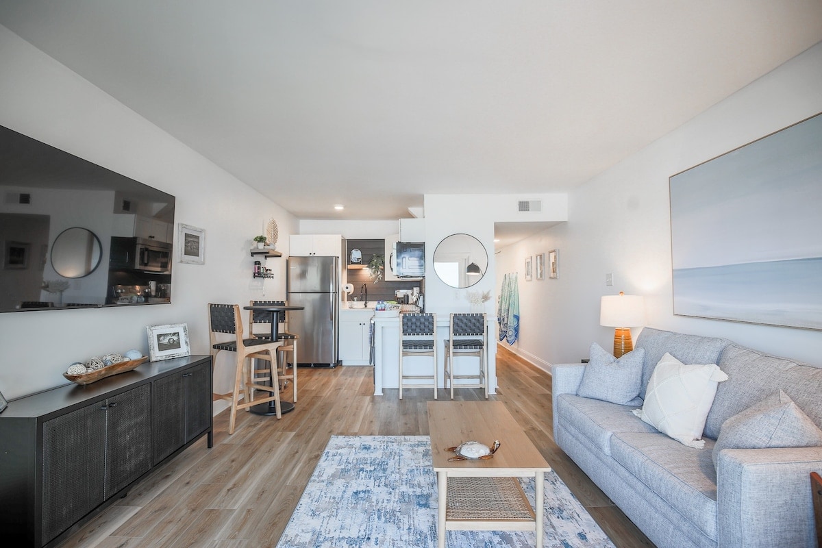 A welcoming living area features a light grey sofa paired with a wooden coffee table atop a blue area rug. A flat-screen TV is mounted on the wall opposite. The dining area includes a round table with four chairs, and the kitchen is visible in the background.