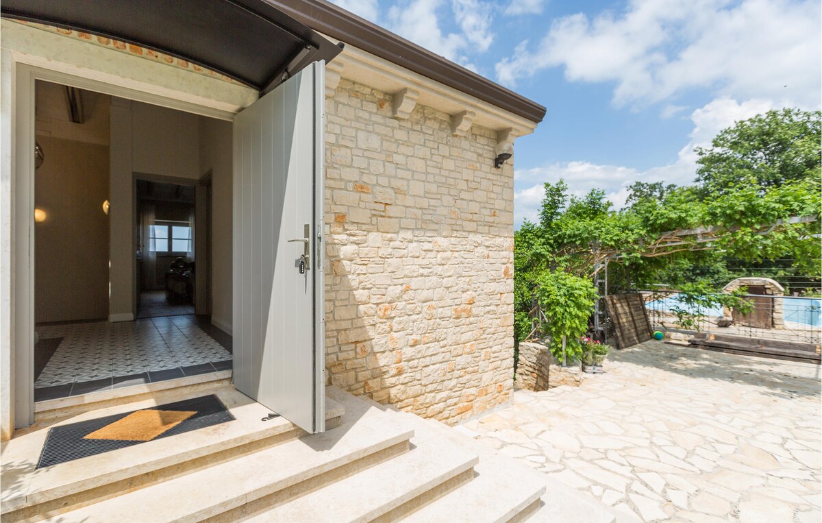 The entrance to the stone house is framed by light-colored stones and features a modern door. Natural light floods the area, revealing a clear view of the garden and large swimming pool beyond the threshold.
