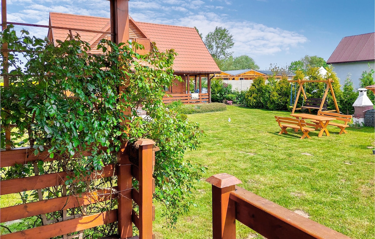 A well-maintained garden is visible, featuring green grass and a wooden swing set. A picnic table is set on one side, complemented by surrounding shrubs and trees. The wooden house stands in the background, with a covered terrace visible, enhancing the natural outdoor setting.