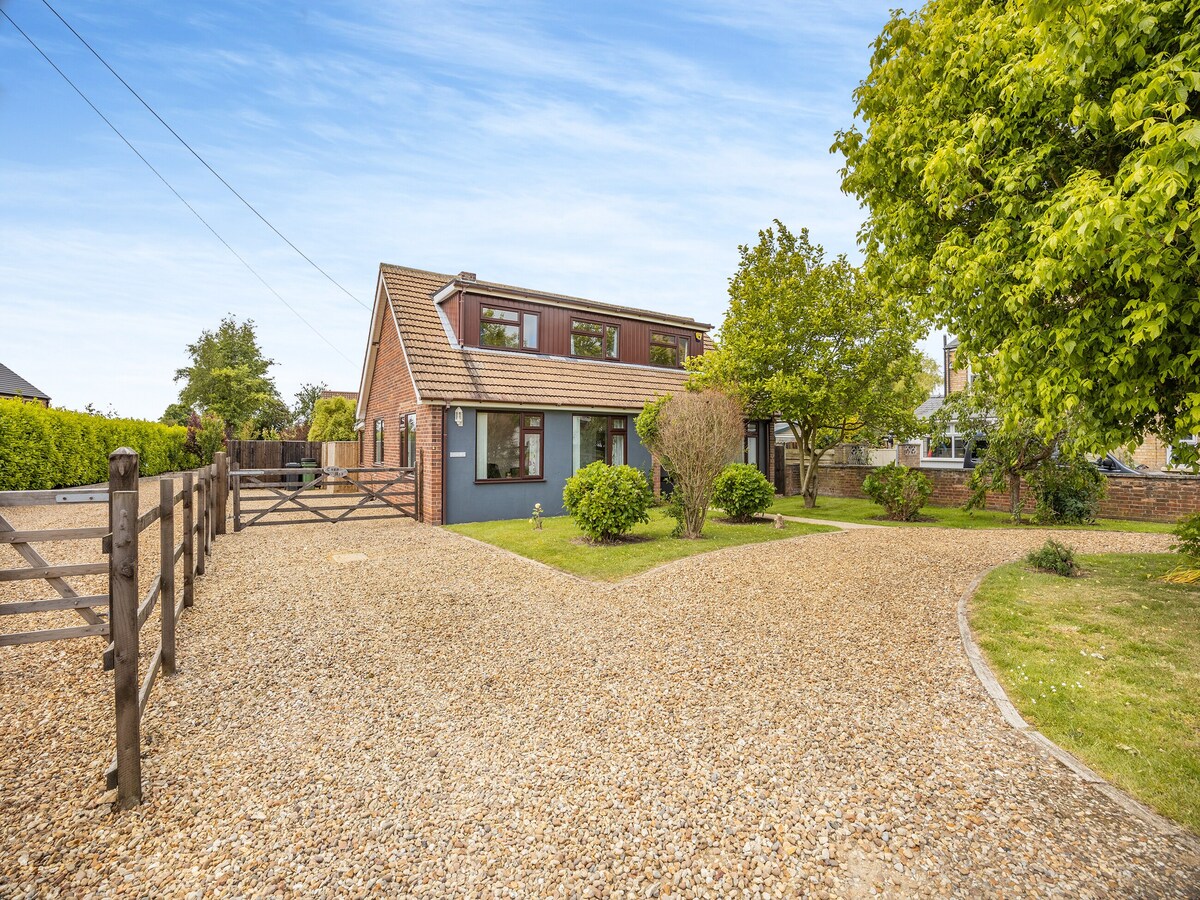 A spacious property is set within a well-maintained garden, featuring a gravel driveway and a combination of shrubs and trees. The two-story home is showcased with a mix of brick and painted exterior, complemented by a lush green backdrop. A welcoming atmosphere is created by the expansive grounds.