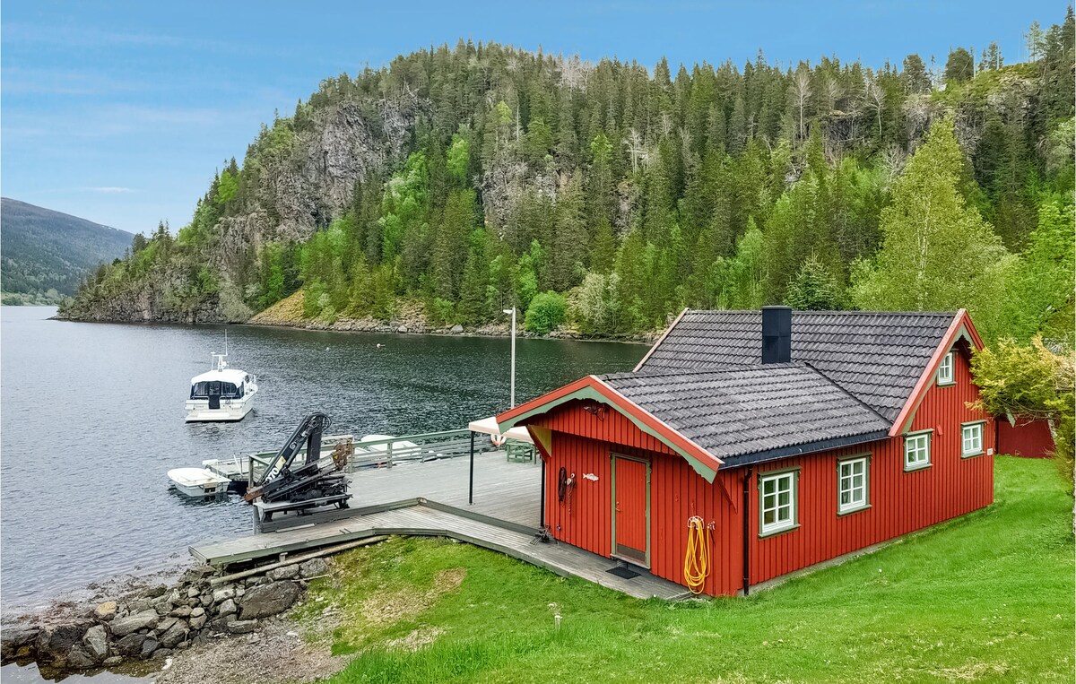 A red-painted wooden cottage is situated near the fjord, with a private pier and a boat moored nearby. Surrounded by lush greenery and rocky cliffs, the area offers a serene natural setting, enhancing the tranquil atmosphere of the location.
