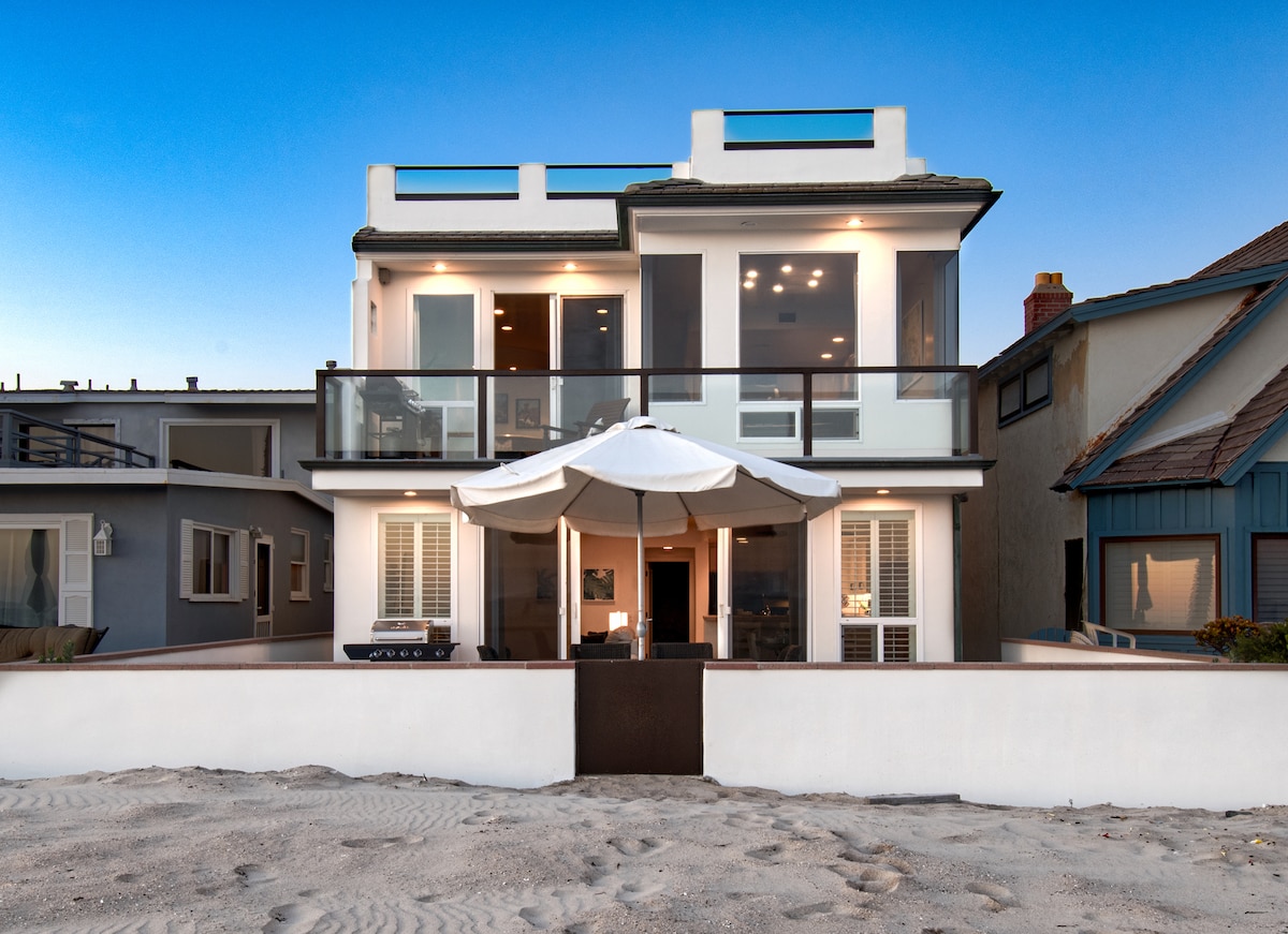 The oceanfront upper unit of a duplex is depicted, featuring large glass doors that provide views of the beach. A broad balcony extends outward, equipped with a large umbrella. The exterior walls are painted in a light color, contrasting with the sandy surroundings.