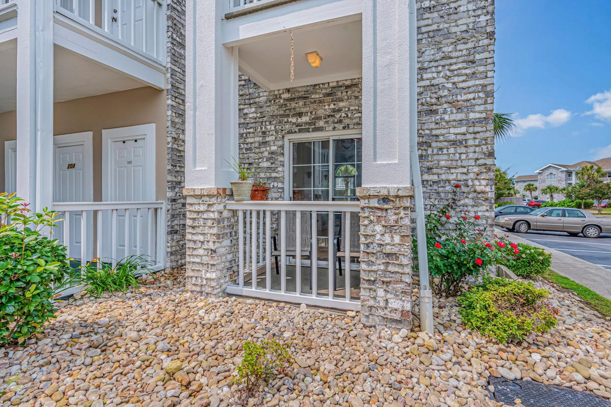 The entrance features a stone pathway leading to an open-air porch with a railing. A small potted plant enhances the welcoming atmosphere. Surrounding greenery and colorful flowers provide natural beauty, while the entrance is clearly marked by a door.