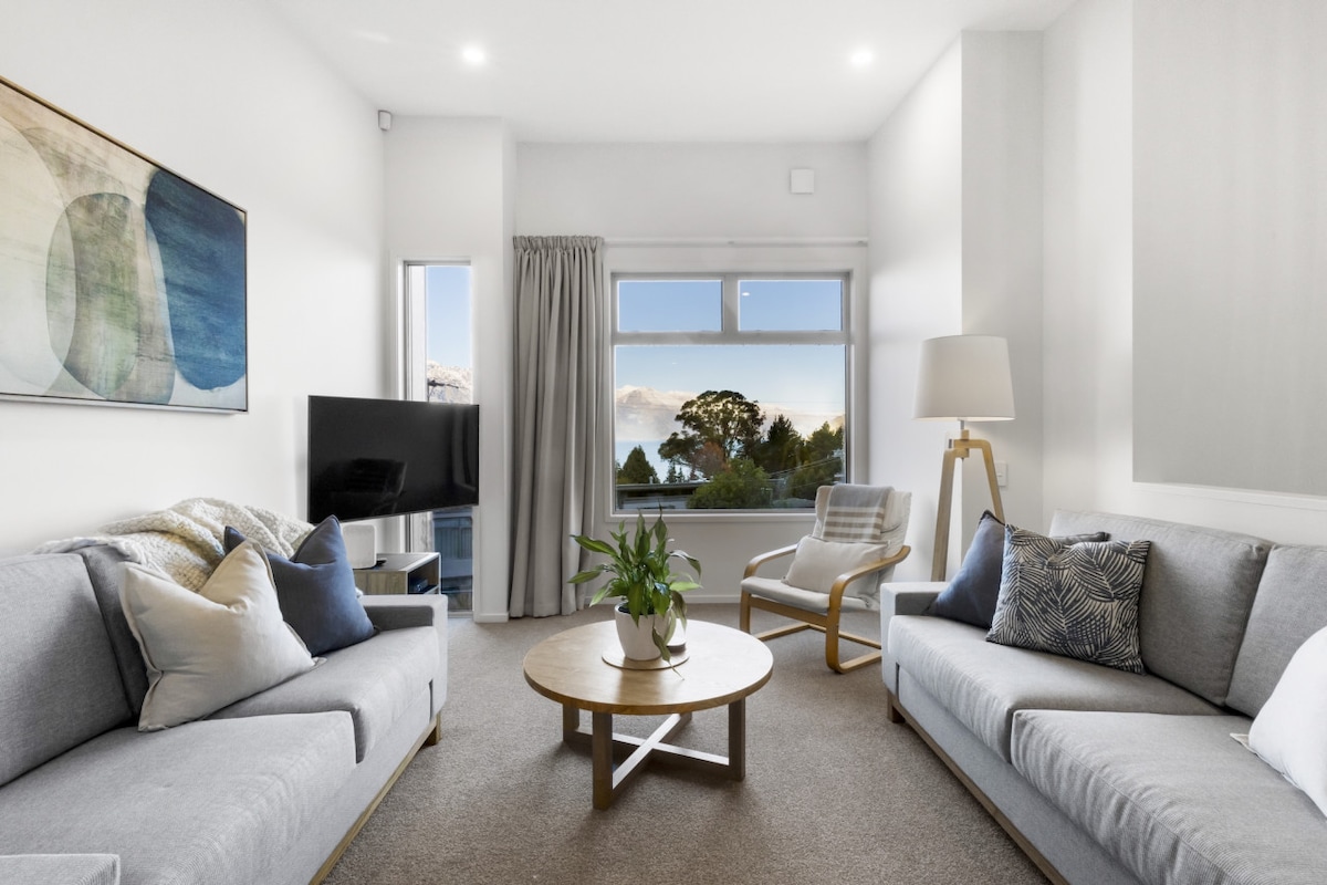 A welcoming living room space features two light gray sofas facing each other. A round coffee table is centered in the room, adorned with a plant. Natural light fills the area from a large window offering lake and mountain views, with a flat-screen TV mounted nearby.