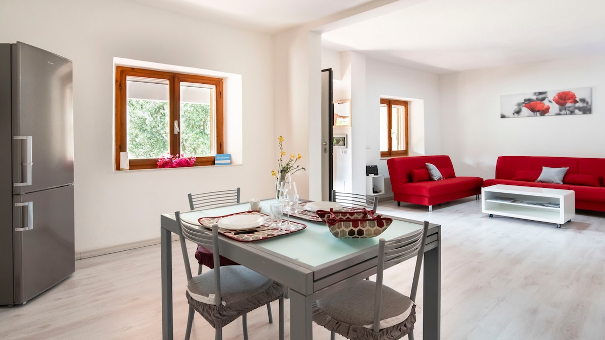 A modern open living space features a dining table set for four, complemented by a contemporary kitchenette and a spacious refrigerator. Two red sofas add a pop of color to the decor, while two windows allow natural light to fill the room.