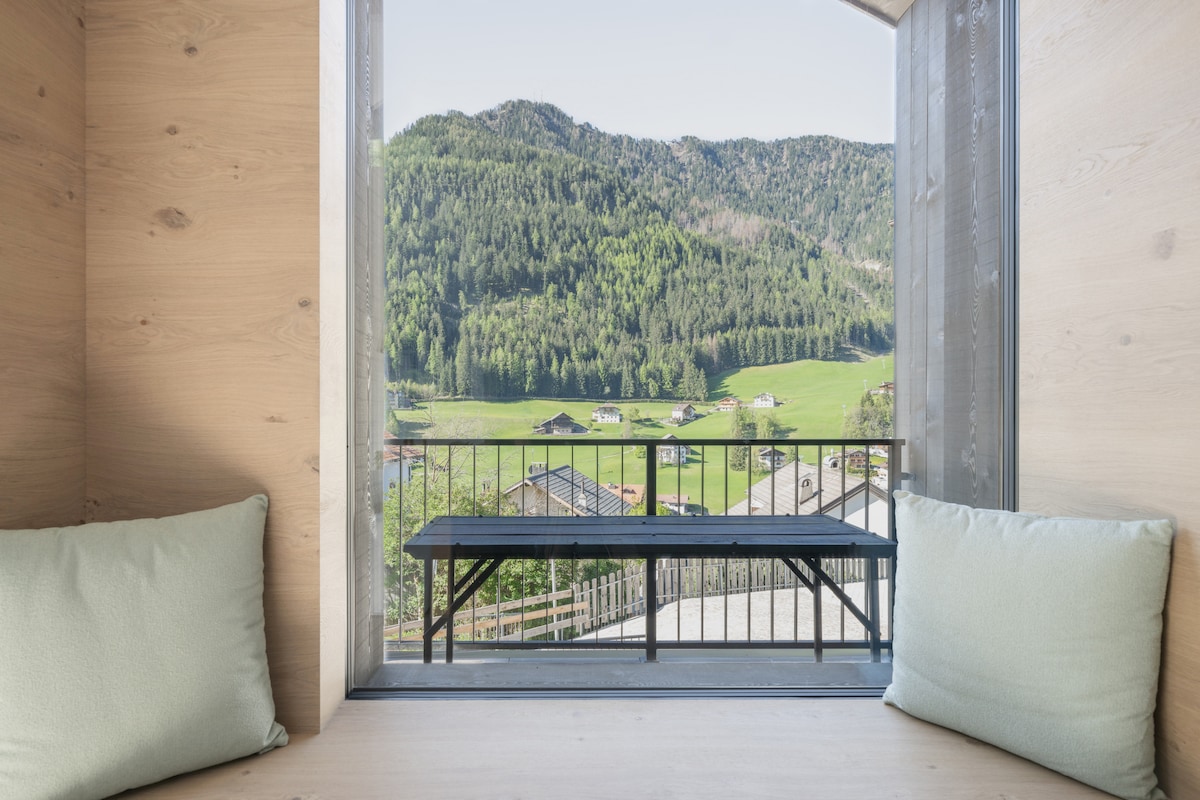 A cozy nook is created by a wide window featuring two soft cushions. The view reveals a lush green landscape with rolling hills and scattered houses under a clear sky.