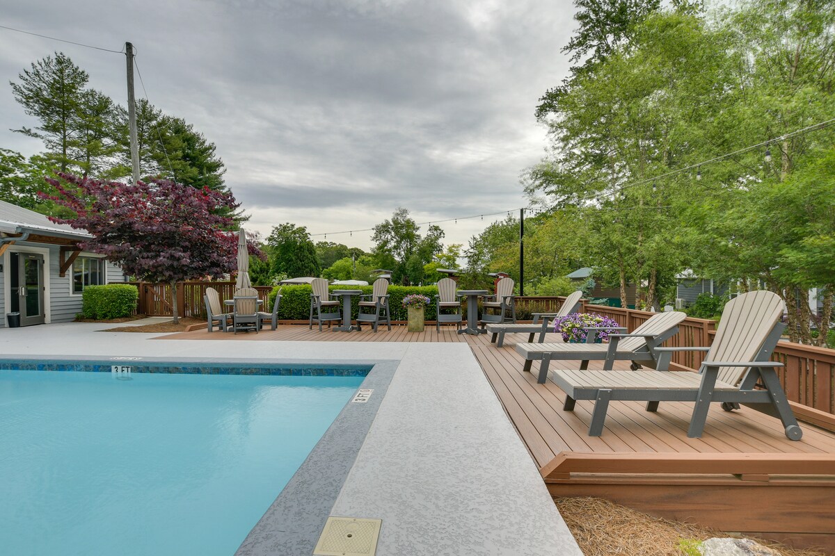 An outdoor pool area is surrounded by wooden decking and lounge chairs. Several tables with chairs are positioned nearby, complemented by trees and shrubs that provide greenery. Soft lighting fixtures are visible above, enhancing the outdoor space.