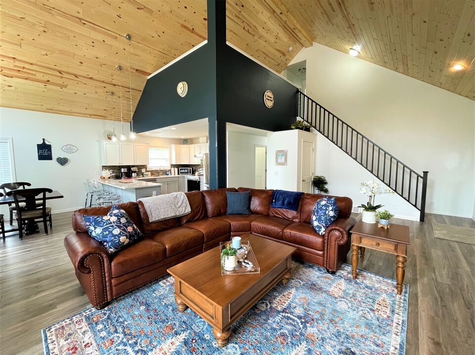 An open living space features a large sectional couch adorned with decorative pillows, situated on a patterned area rug. A wooden coffee table rests in front of the couch, and a dining area with a round table is visible in the background. Cathedral ceilings enhance the openness.
