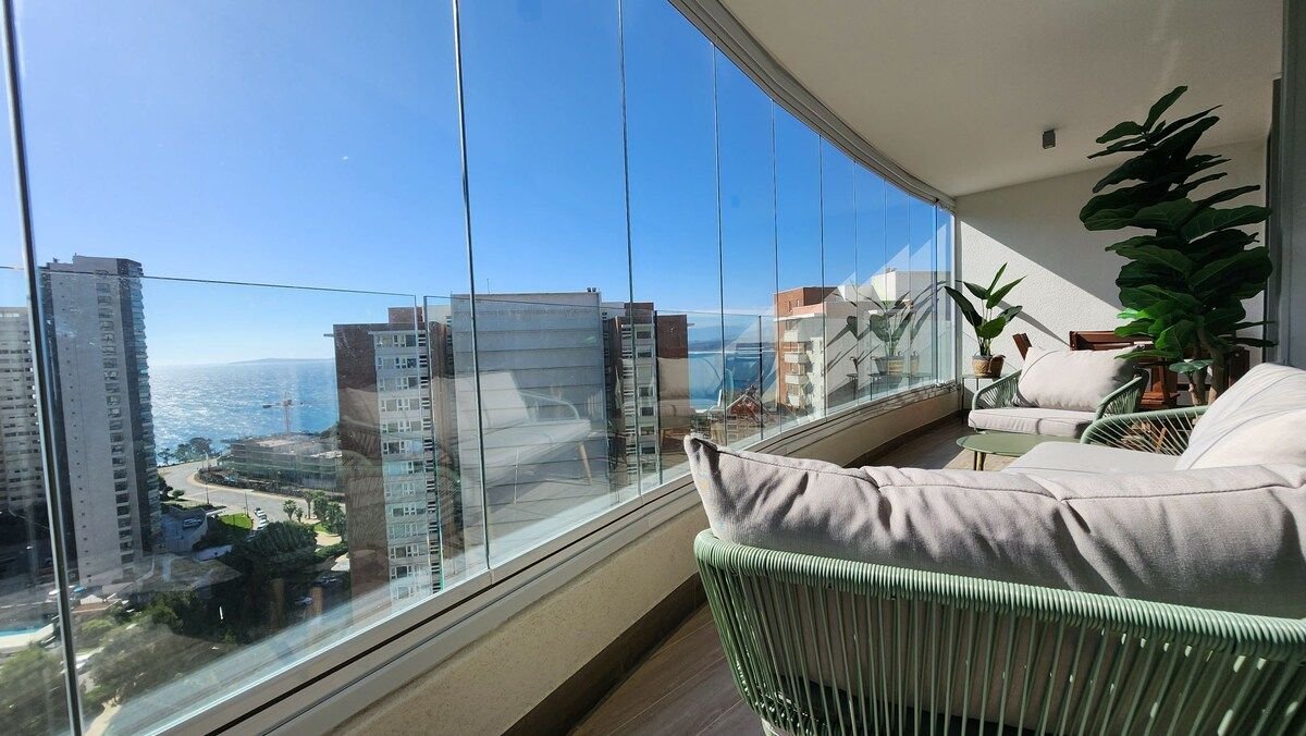 A bright and airy lounge area features floor-to-ceiling windows that provide a panoramic view of the ocean and surrounding buildings. Comfortable seating is arranged with plush cushions, complemented by potted greenery, adding a touch of nature to the inviting space.