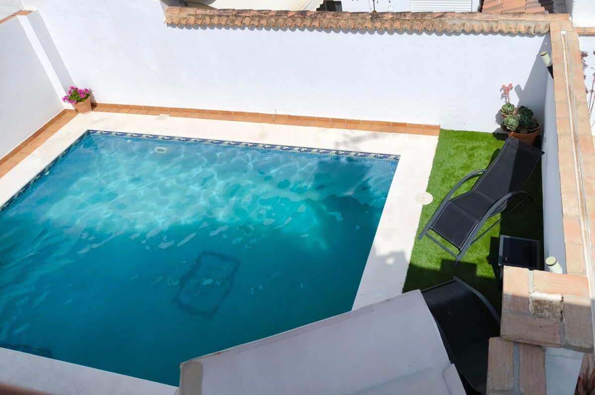 A private pool sits within a walled area, surrounded by whitewashed walls and decorative plants. Two sun loungers are positioned on grass-like turf beside the pool, which reflects clear blue water under sunlight.