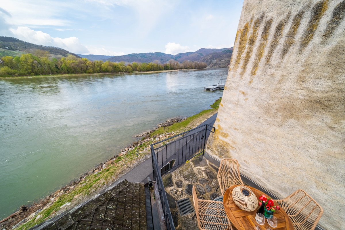 A small outdoor seating area features a round wooden table accompanied by two rattan chairs. Scenic river views are presented, with lush greenery lining the opposite bank and distant hills under a partly cloudy sky.