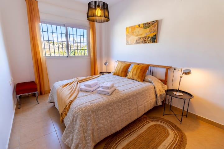 ห้องนอนพร้อมเตียงคู่ ผ้าคลุมเตียงสีอ่อน หมอนสีเหลืองอ่อน ผ้าห่มและผ้าเช็ดตัว มีโต๊ะ 2 ตัวพร้อมโคมไฟ ตู้เสื้อผ้าในตัวที่กว้างขวาง เครื่องปรับอากาศ หน้าต่างพร้อมตาข่ายและผ้าม่าน พรมยูท และโคมไฟเพดานสีดำ