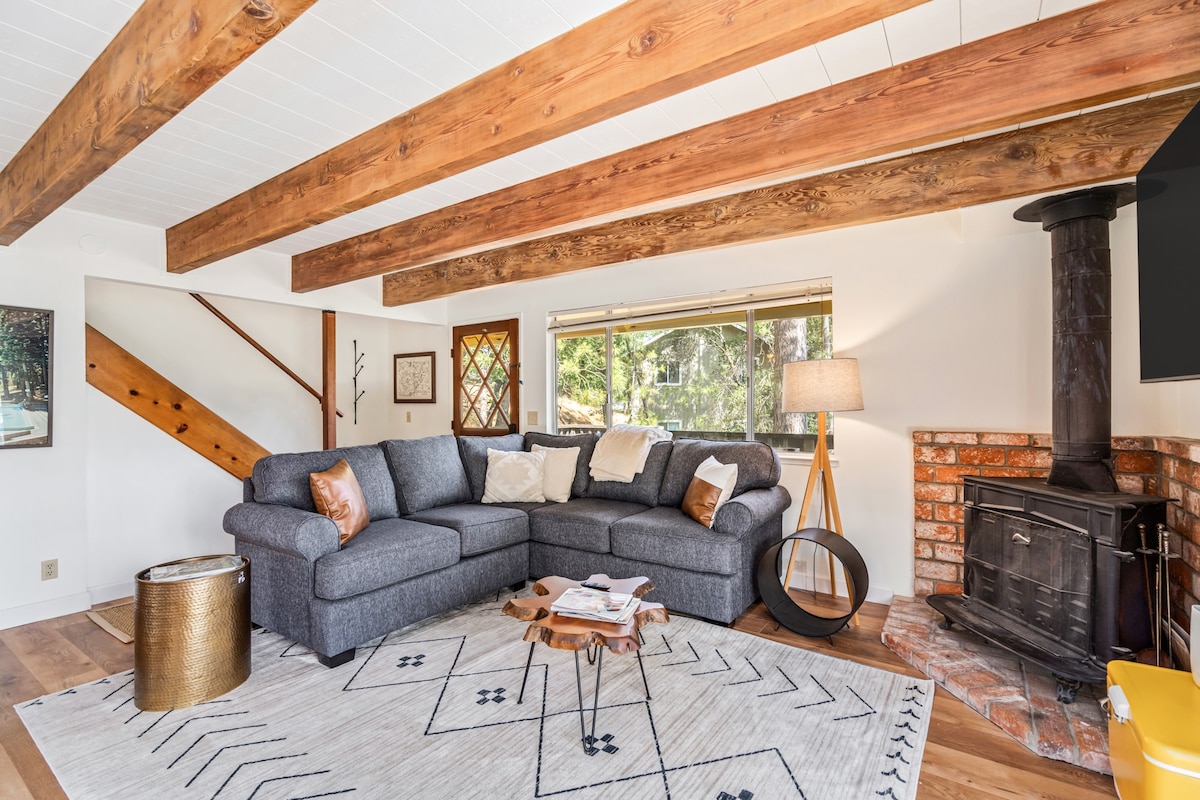 A spacious living area features a large sectional sofa positioned on a light rug, accompanied by a round coffee table. Exposed wooden beams draw attention overhead, while a brick fireplace adds warmth to the room. Natural light filters through a wide window, creating an inviting space.