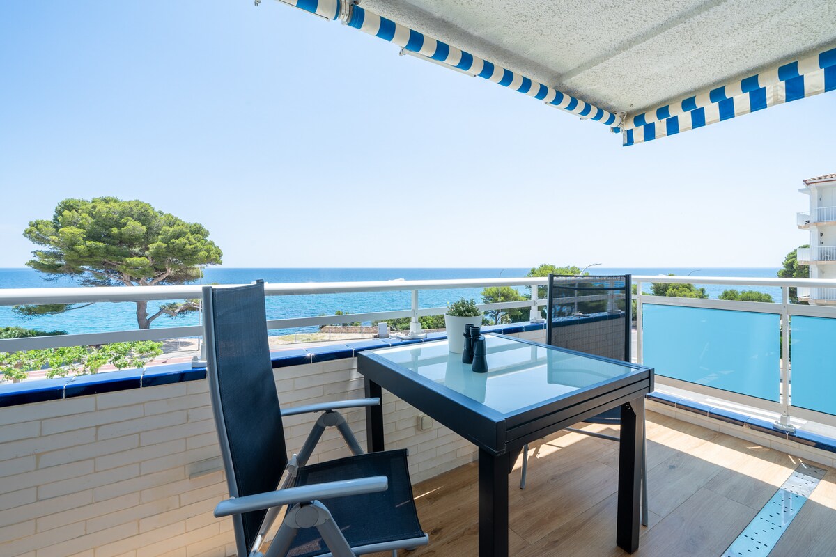 A private covered terrace features a glass-top table with seating for four, offering views of the Mediterranean Sea. The space is complemented by a sunshade and surrounded by a railing, providing an open yet sheltered area for relaxation.