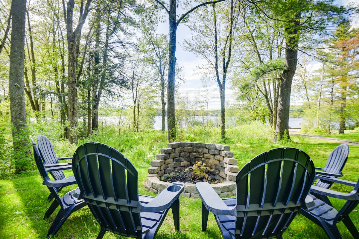 A fire pit encircled by several dark blue Adirondack chairs is situated on a grassy area. The surrounding landscape features lush greenery and trees, with a tranquil pond visible in the background under a clear sky.