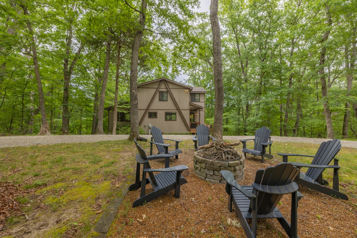 An outdoor fire pit area is surrounded by several black adirondack chairs, set on a gravel surface. Lush greenery and mature trees provide a serene backdrop to the modern a-frame chalet visible in the distance.