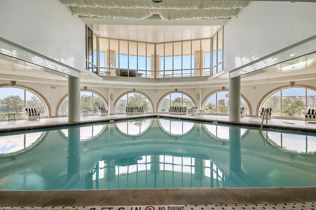 An indoor pool area features large arched windows, allowing natural light to flood the space. The calm water reflects the surroundings, while lounge chairs are arranged nearby for relaxation. Decorative tiles line the pool's edge, ensuring a clean, inviting environment.