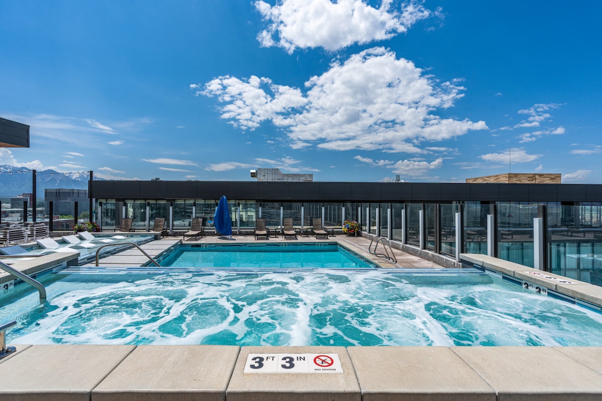 A rooftop pool and hot tub are featured in an expansive area with panoramic views of the sky and mountains. Lounge chairs are arranged around the pool, and a safety sign is positioned near the edge of the hot tub, indicating a maximum depth.