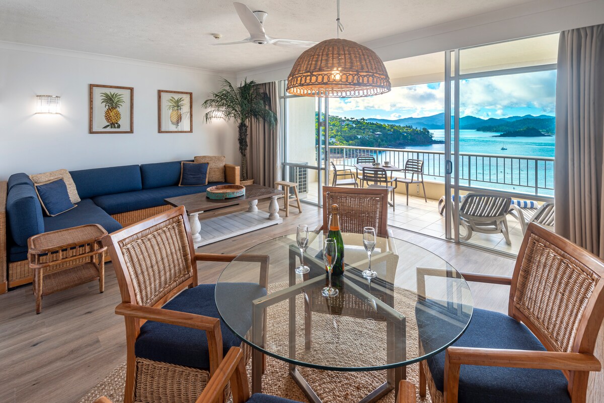 A spacious living area is highlighted with a modern sectional sofa and a round glass dining table surrounded by wooden chairs. Natural light floods the space through large sliding doors, which open to a balcony with scenic views of the water and mountains.