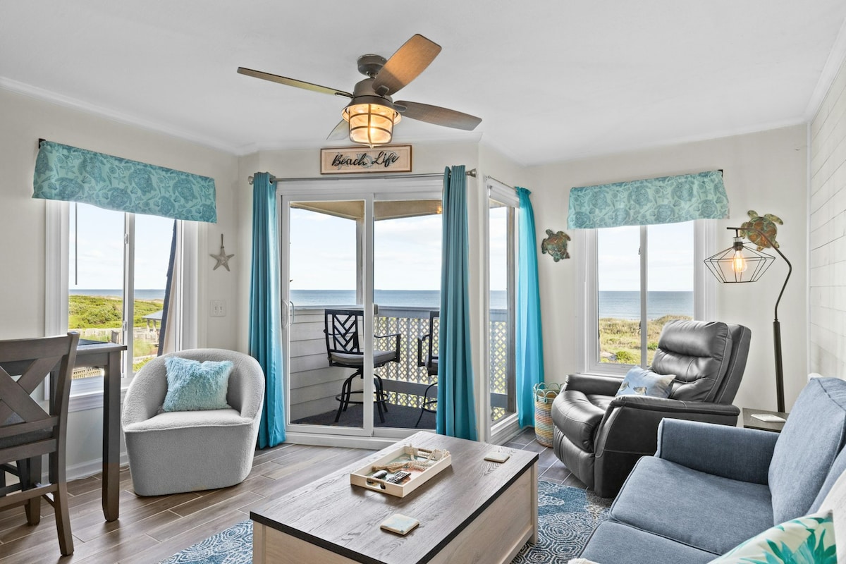 The living area features a comfortable sofa paired with a recliner, with large windows allowing natural light and ocean views. A wooden coffee table sits in the center, and a dining table is positioned nearby. Breezy blue curtains frame the windows, while ceiling fan blades circulate air.