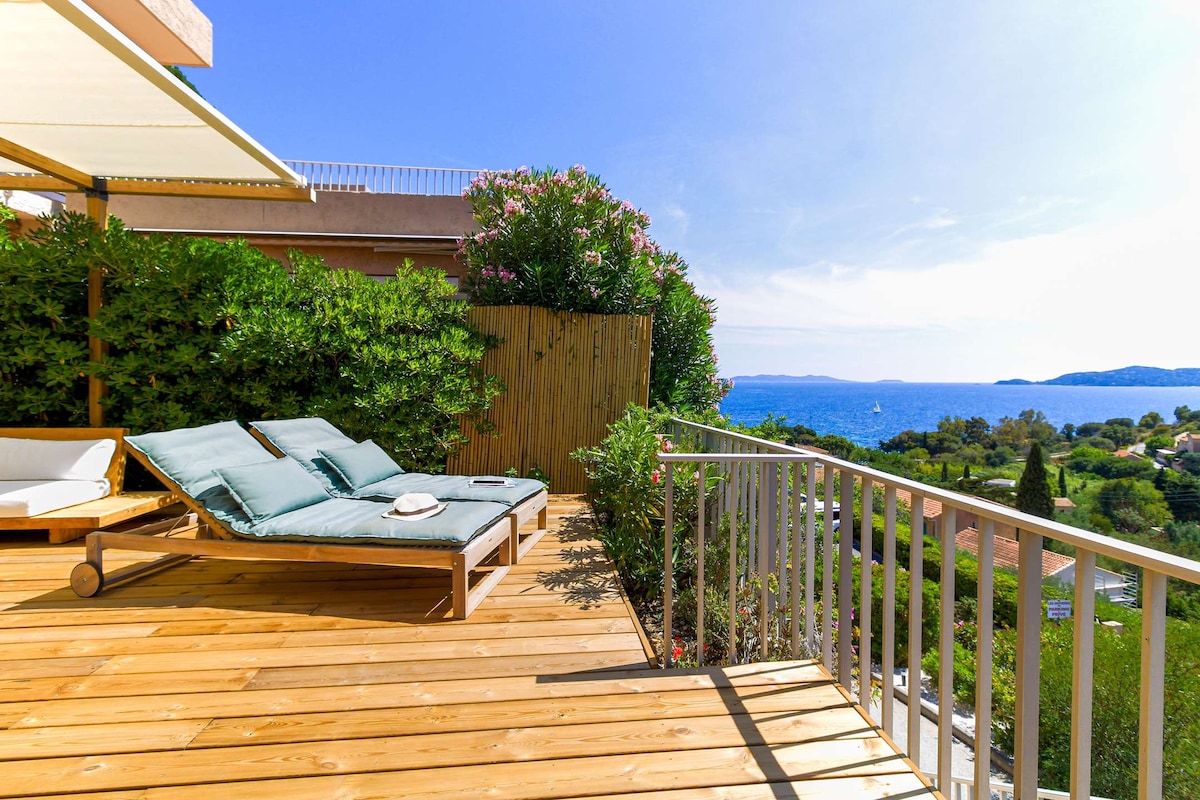 A spacious wooden terrace features a pair of sun loungers positioned for relaxation and ocean views. Lush greenery surrounds the area, enhancing privacy. The clear blue sky complements the tranquil sea in the background, creating a serene outdoor environment.