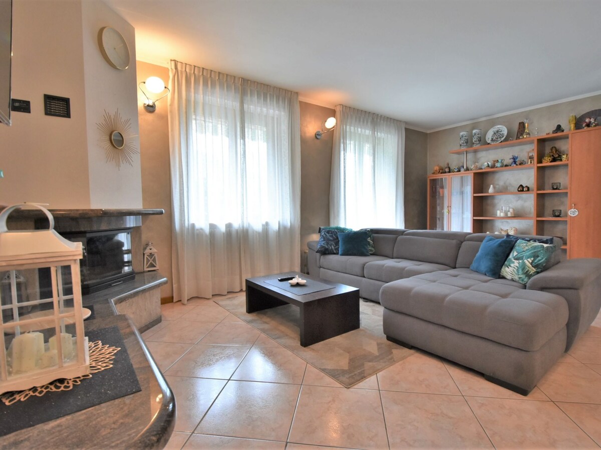 A spacious living room is highlighted by large windows dressed in sheer curtains, allowing natural light to fill the space. A modern sectional sofa is complemented by decorative cushions. A coffee table sits in front, with a cozy corner featuring a fireplace and decorative shelves.