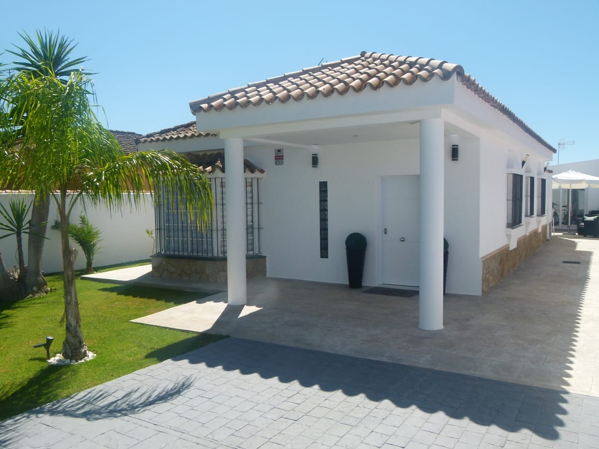 The exterior of the holiday home features a white façade with a terracotta tiled roof. A manicured lawn with palm trees borders the entrance, which is framed by two tall planters. The driveway is paved, providing easy access to the property.