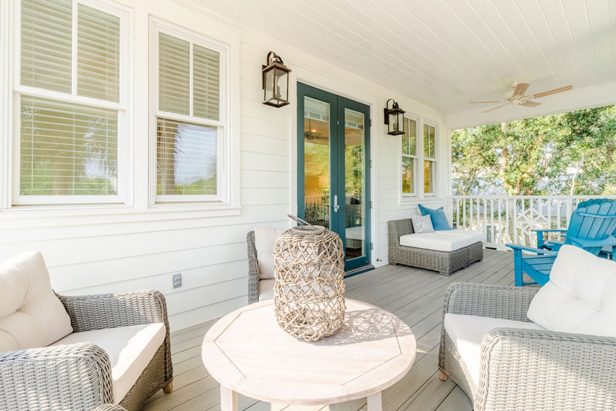 A spacious front porch is depicted, featuring comfortable seating arrangements with light-colored cushions and a round wooden table at the center. Large windows provide an inviting view, while decorative lanterns hang from the walls, contributing to a relaxing outdoor space.