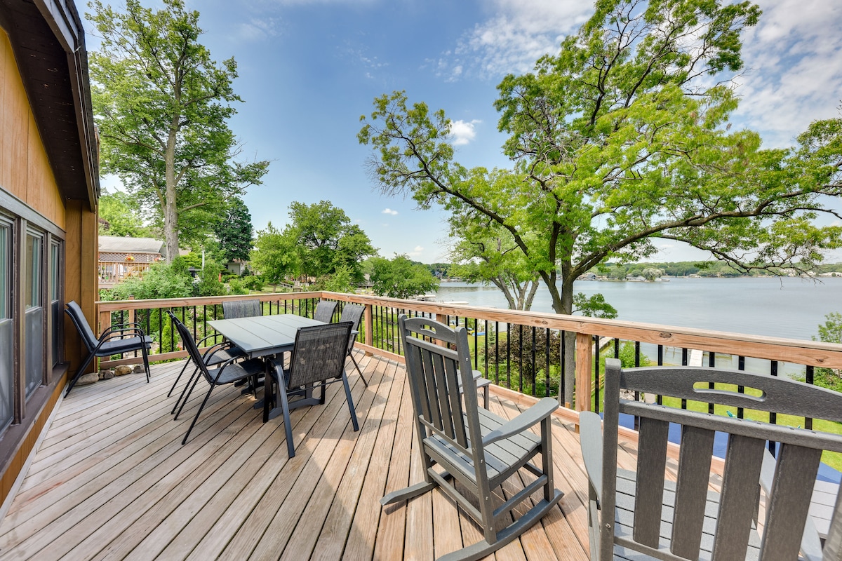 A wooden deck extends over tranquil waters, featuring a dining table surrounded by several chairs. Adirondack chairs are positioned nearby, and lush greenery frames the scene, providing a serene outdoor space for relaxation and enjoyment.