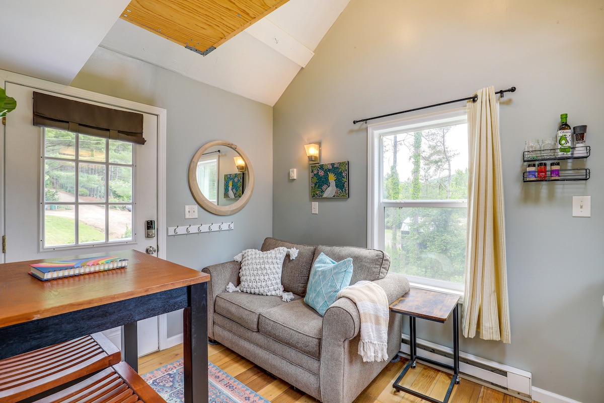 A cozy corner of the studio features a comfortable sofa adorned with decorative pillows. Natural light fills the space through a large window, while a small side table complements a compact dining area. Shelves display thoughtfully arranged items, adding a personal touch to the room.