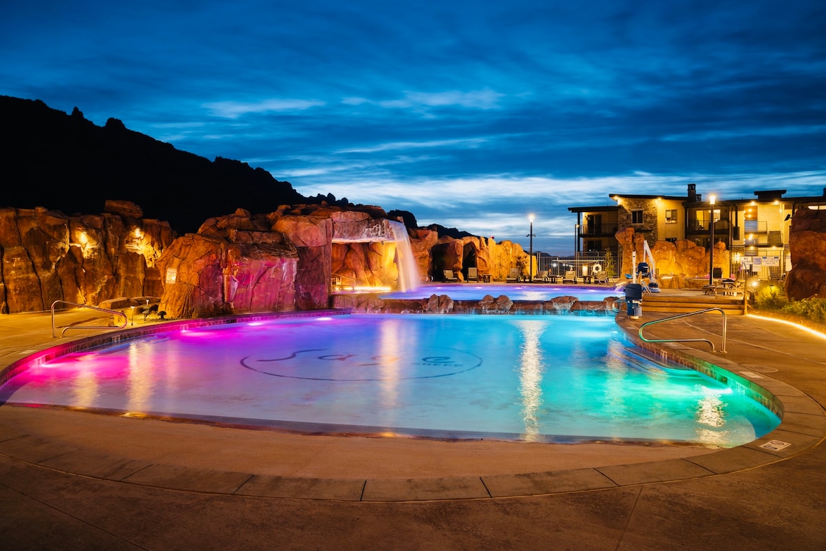 A serene pool area is illuminated by colorful lights, creating a calming ambiance. Surrounding rock formations and a waterfall enhance the natural setting, while lounge chairs line the pool's edge. The evening sky transitions from blue to dusk, contributing to a relaxing atmosphere.