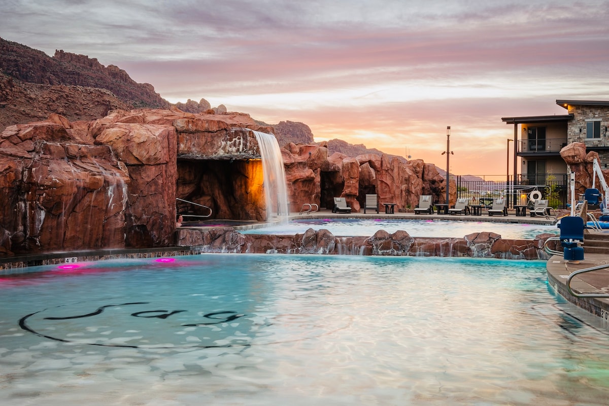 A resort-style pool area is featured, highlighted by a waterfall cascading over rocky formations. Vibrant lighting enhances the water's surface. Lounge chairs are arranged around the pool, providing seating options. The background showcases the complex's modern architecture and the surrounding red cliffs at sunset.