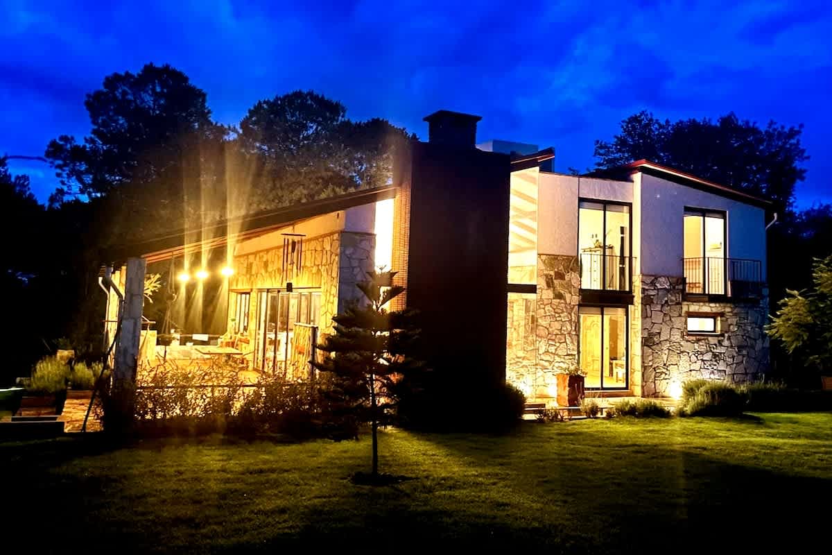 An elegant house is illuminated against a darkening sky, showcasing a combination of stone and modern architecture. Warm light spills from large windows, highlighting the inviting outdoor space. A well-kept lawn complements the structure, surrounded by trees creating a serene atmosphere.