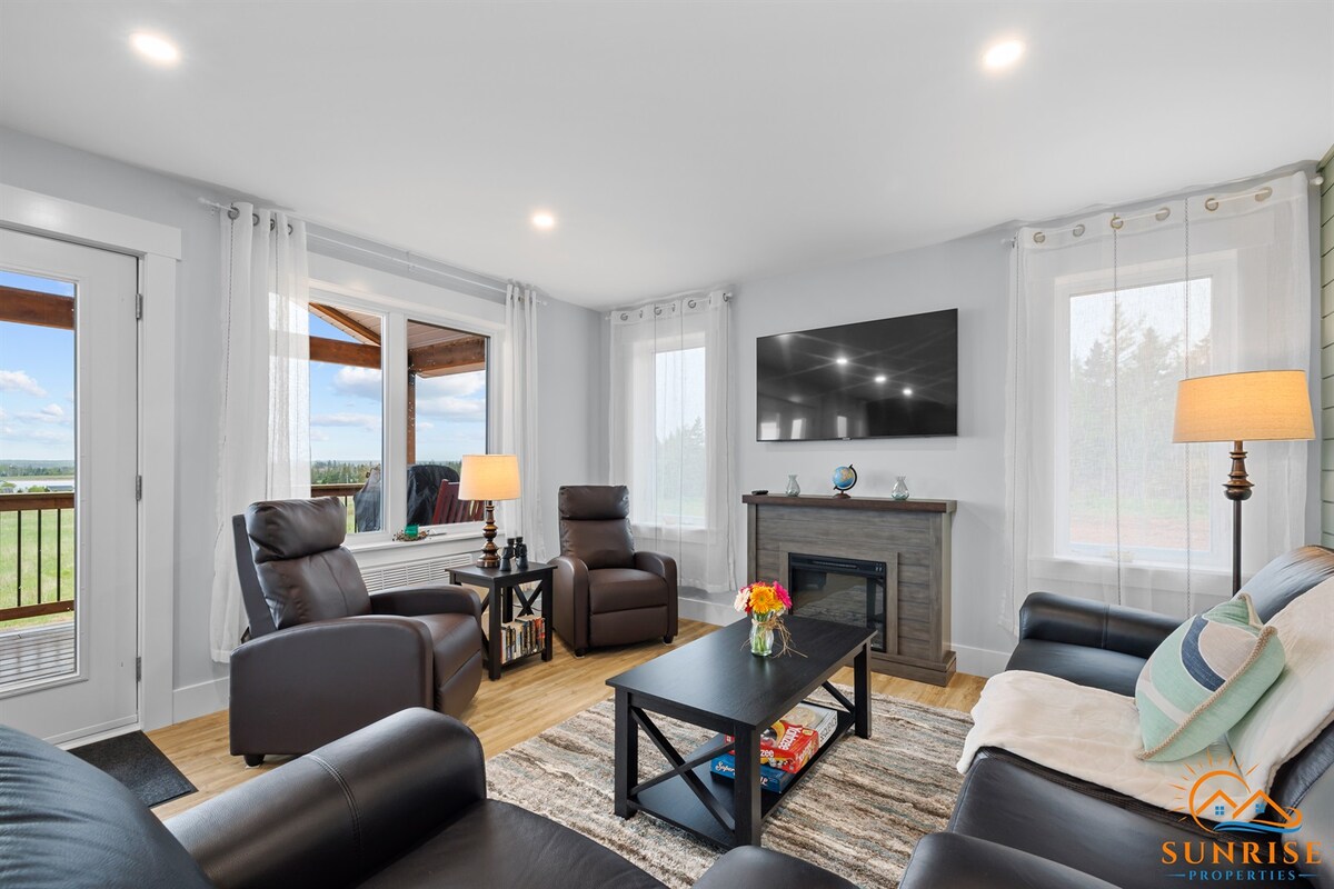 A spacious living room is visible, featuring comfortable seating arrangements, including leather recliners and a sofa. Natural light floods the area through sheer curtain-covered windows. A modern flat-screen television is mounted on the wall, with a tasteful coffee table positioned in front, complemented by a decorative vase.