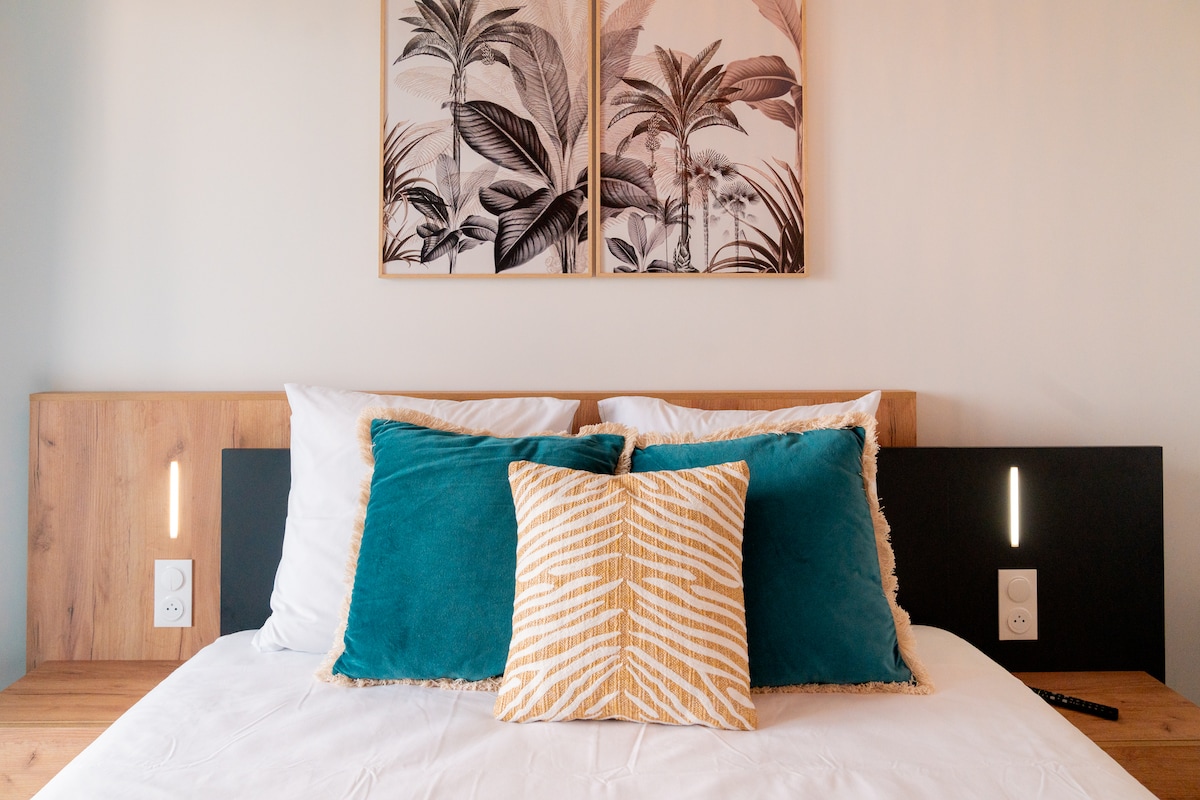 A spacious bed is featured in the sleeping area, adorned with multiple decorative pillows in turquoise and gold. Two light fixtures are integrated into the wooden headboard, providing a modern touch. Framed botanical art hangs above the bed, adding a decorative element to the room.