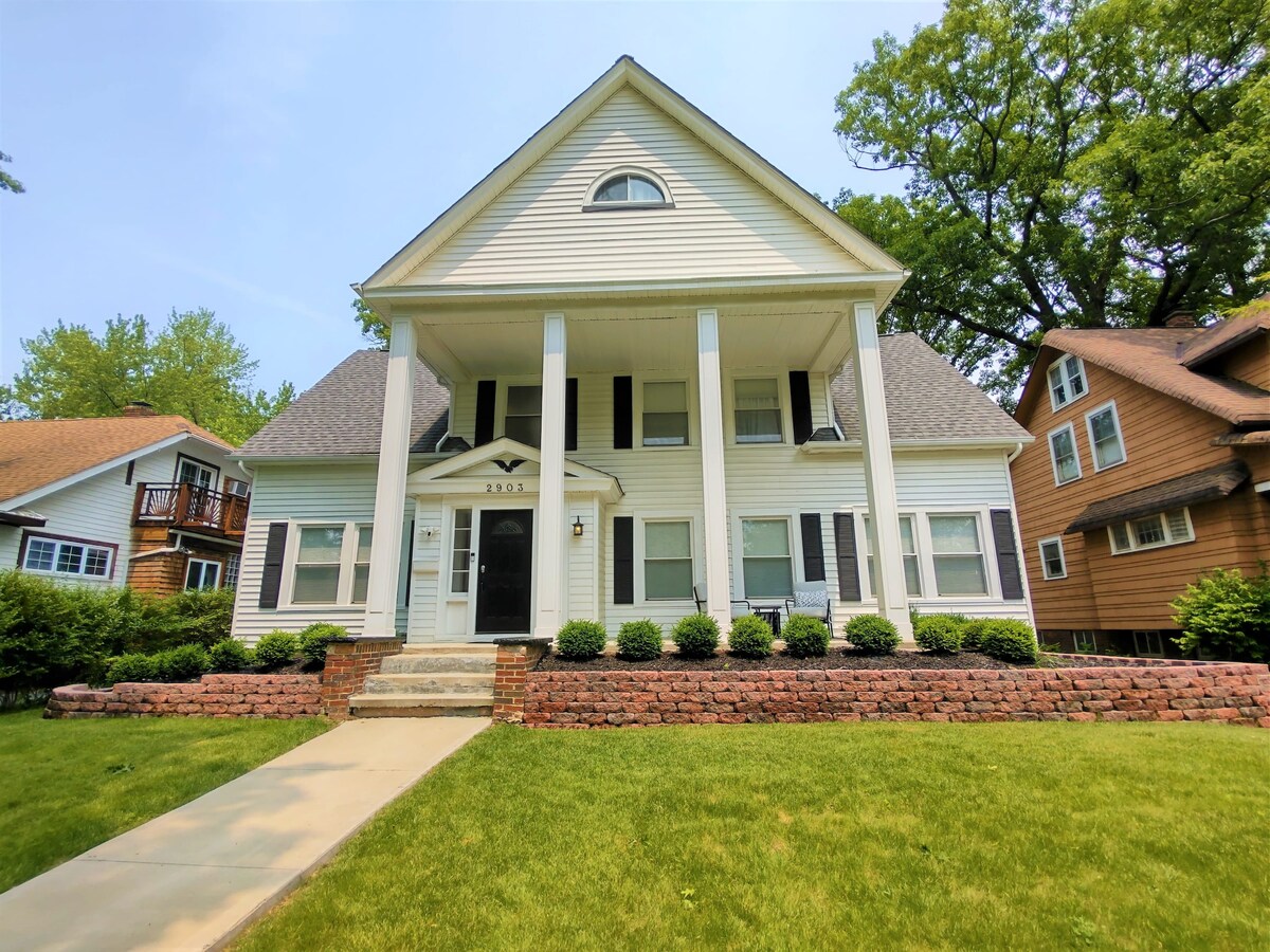 A large, two-story Colonial home is depicted with a welcoming façade. The exterior features a light-colored siding, large porch with white columns, and trimmed bushes along the front. A well-manicured lawn creates a pleasant entrance, and trees provide shade in the background.