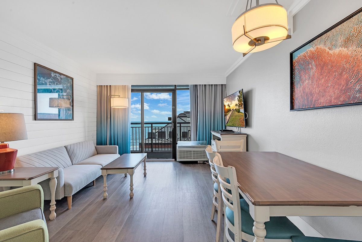A light and airy living area features a comfortable seating arrangement, including a gray sofa and two accent chairs. A wooden dining table with four chairs is positioned nearby. Large sliding doors lead to a balcony, allowing natural light to fill the space.