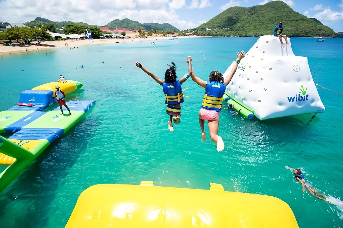Two individuals in life vests leap off a floating platform into clear blue water, surrounded by inflatable water attractions. In the background, a sandy beach and greenery are visible, alongside a mountainous landscape under a bright sky.