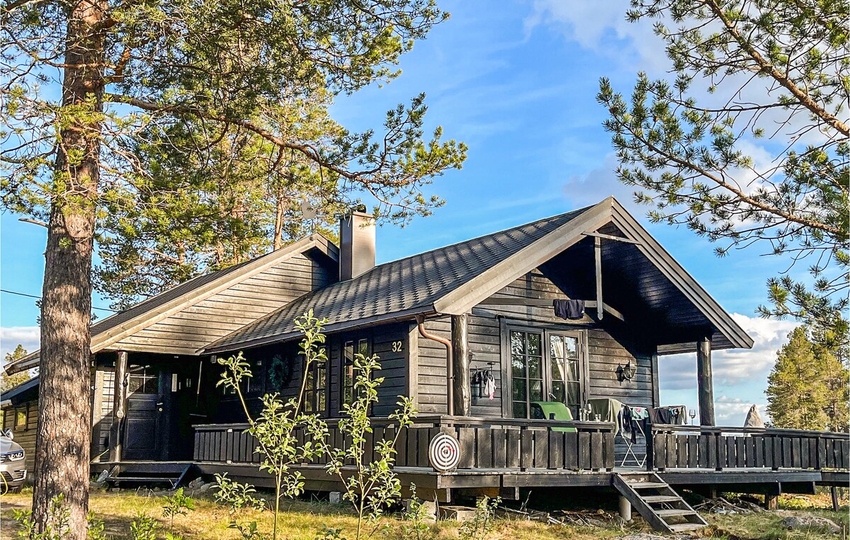 A charming mountain cottage features a dark wooden exterior and a steep roof. The inviting porch extends towards a grassy area, bordered by tall trees. Large windows allow natural light to fill the space, while comfortable seating is visible, offering opportunities for relaxation and enjoyment of the outdoors.
