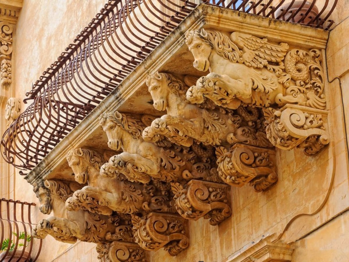 An elaborate balcony features intricately carved horse sculptures serving as decorative corbels. The detailed craftsmanship highlights swirling patterns and ornate motifs. A wrought-iron railing complements the historical architecture, enhancing the elegance of the exterior.