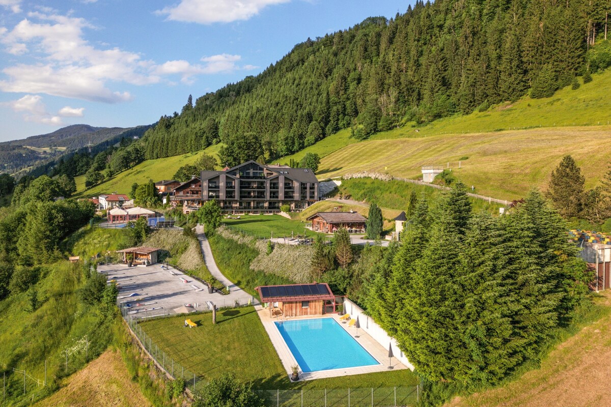A panoramic view of the Weitenmoos Panorama Apartments set against a backdrop of rolling green hills. The lush landscape features a large outdoor pool surrounded by sun loungers and shaded areas. Nearby, wooden structures and recreational spaces are nestled within the natural surroundings.
