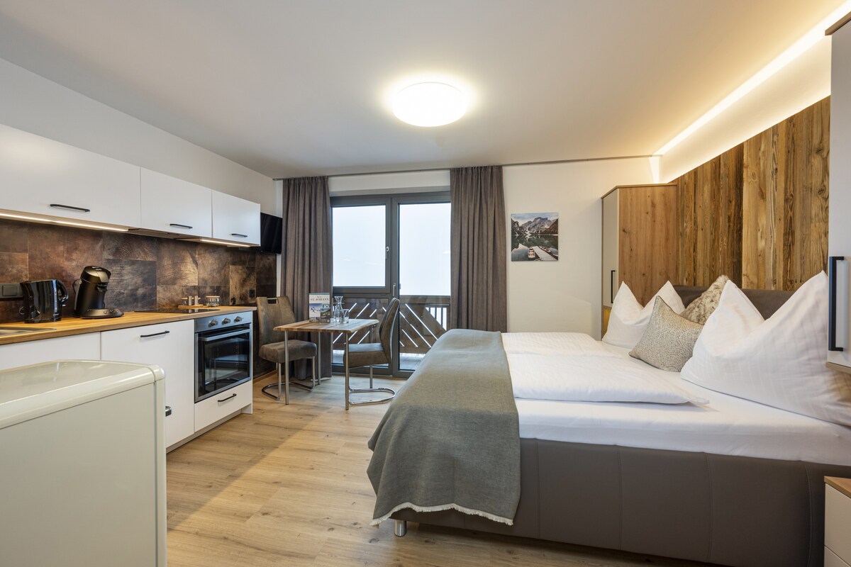 A modern studio features a combined living and sleeping area with a comfortable double bed and a small seating space. The fully equipped kitchenette includes a refrigerator, oven, and coffee machine. Large windows allow natural light to flow in, and access to a balcony is provided.