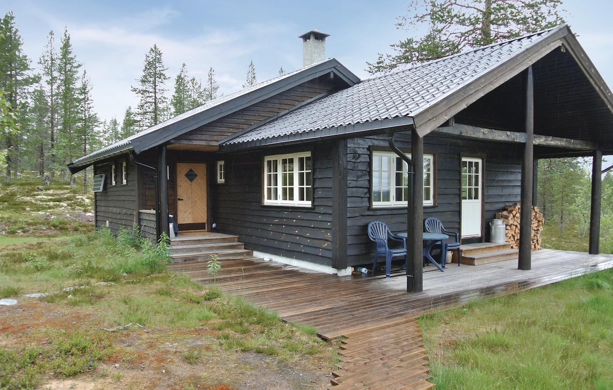 The cottage features a dark wooden exterior with a sloped roof and a welcoming porch. Two chairs are placed on the covered deck, and firewood is stacked beside the entrance. Surrounding greenery includes low shrubbery and scattered trees, adding to the natural setting.